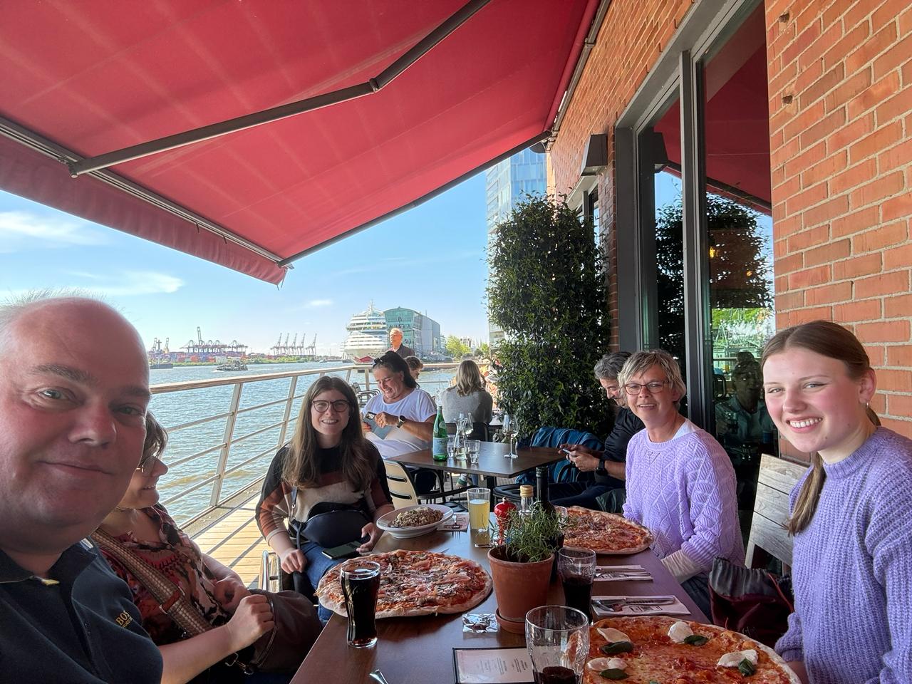 Sechs Rollifahrer essen bei strahlend blauem Himmel Pizza am Hamburger Hafen.